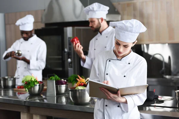 Female chef writing in recipe book by her multiracial colleagues — Stock Photo