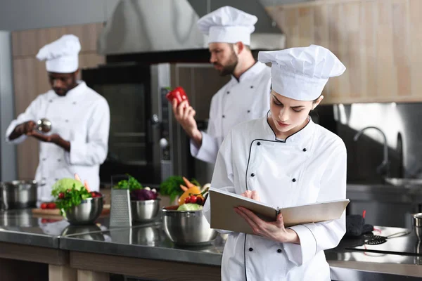 Femme chef écrivant dans un livre de recettes par ses collègues multiraciaux — Photo de stock