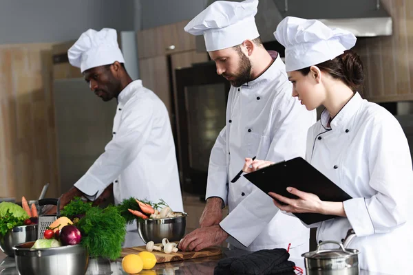 Female chef writing down recipe by her multiracial colleagues — Stock Photo