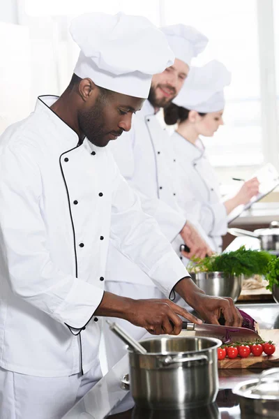 Équipe de chefs multiraciaux coupant des ingrédients par table de cuisine — Photo de stock