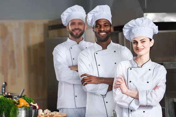 Équipe de chefs multiraciaux posant les bras croisés sur la cuisine — Photo de stock