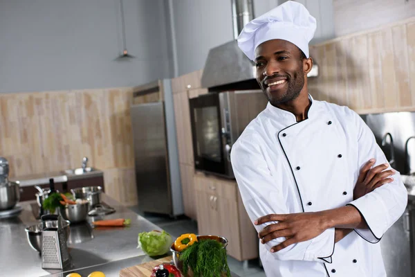 Chef afroamericano de pie con los brazos cruzados por la mesa con ingredientes de cocina - foto de stock