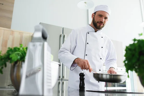 Low angle view of handsome chef frying vegetables on frying pan at restaurant kitchen — Stock Photo