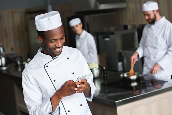 Un chef afro-américain souriant utilisant un smartphone dans la cuisine du restaurant — Photo de stock