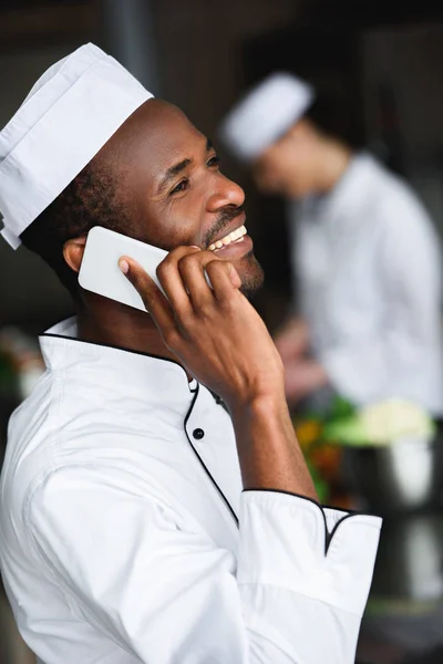 Vista lateral del sonriente chef afroamericano hablando por teléfono inteligente en la cocina del restaurante — Stock Photo