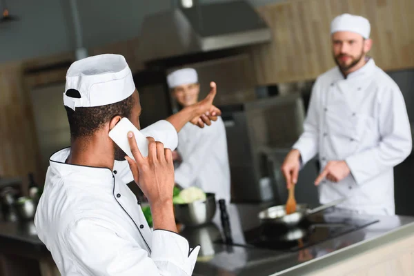 Chef afro-américain parlant par smartphone et montrant pouce à ses collègues à la cuisine du restaurant — Photo de stock