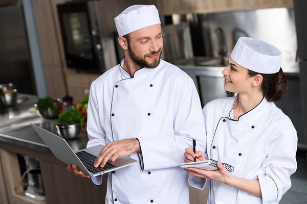 Chefs using laptop at restaurant kitchen and looking at each other — Stock Photo