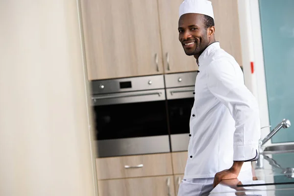 Vista lateral del guapo chef afroamericano apoyado en el mostrador de la cocina en la cocina del restaurante - foto de stock