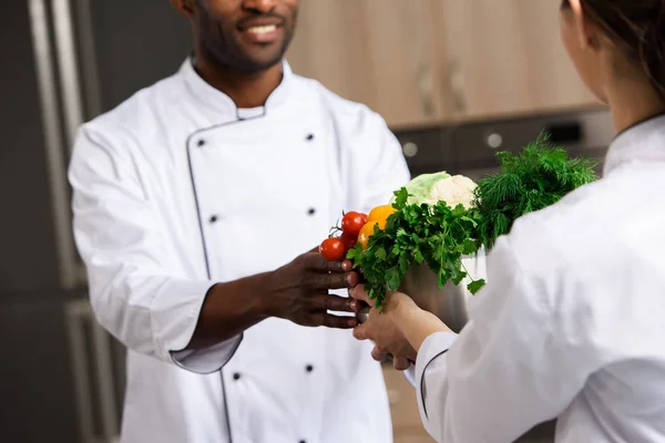 Imagen recortada de chef dando tazón con verduras a un colega afroamericano en la cocina del restaurante - foto de stock