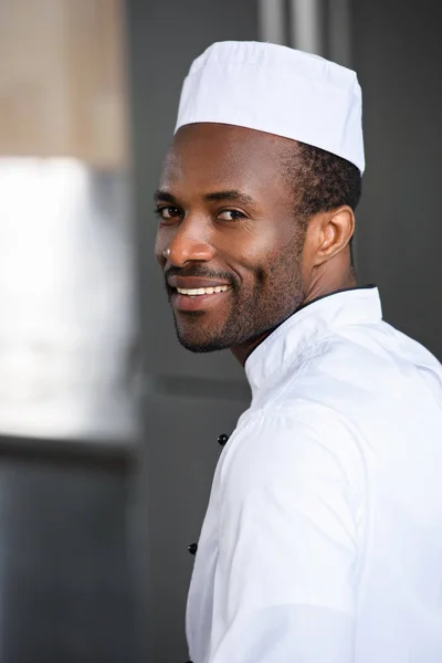 Retrato del guapo y sonriente chef afroamericano mirando a la cámara en la cocina del restaurante - foto de stock