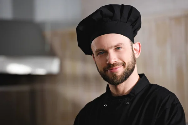 Portrait de beau chef en uniforme noir regardant la caméra à la cuisine du restaurant — Photo de stock
