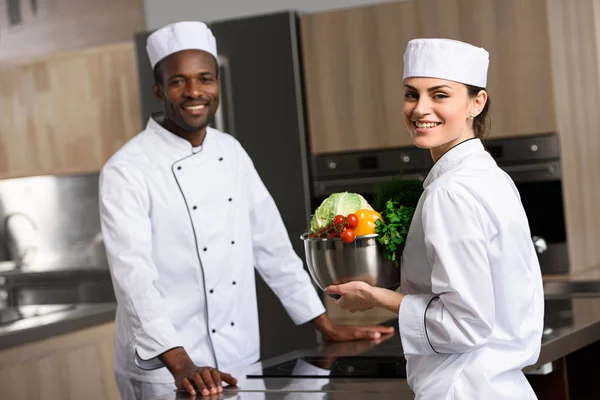 Multicultural chefs holding vegetables and looking at camera at restaurant kitchen — Stock Photo