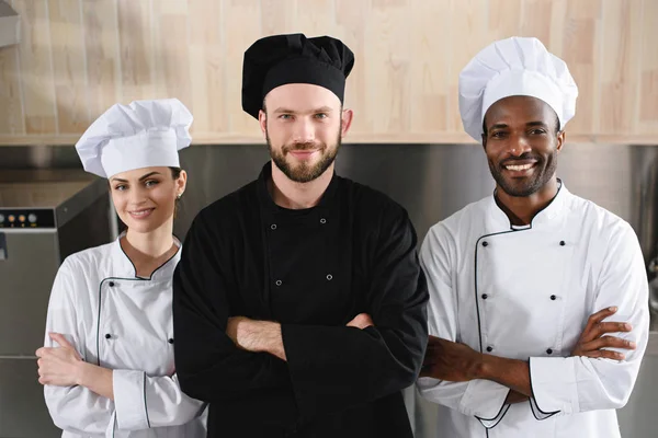 Multicultural chefs standing with crossed arms and looking at camera at restaurant kitchen — Stock Photo