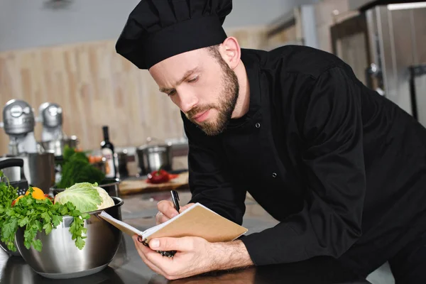 Seitenansicht des gutaussehenden Kochs, der in der Restaurantküche neue Rezepte auf Notizbuch schreibt — Stockfoto