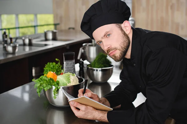 Handsome chef writing down new recipe to notebook at restaurant kitchen and looking away — Stock Photo