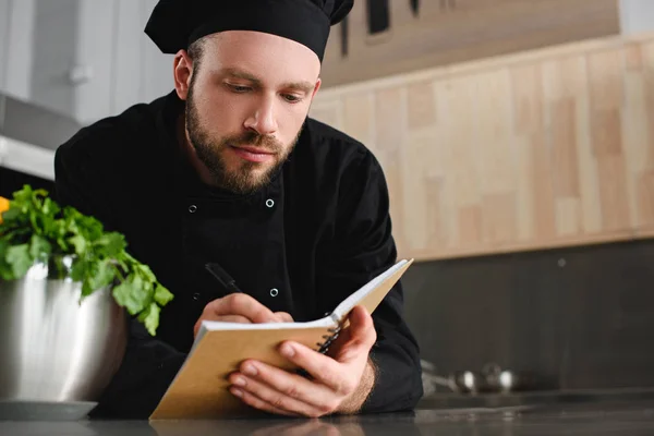 Handsome chef writing down new recipe to notebook at restaurant kitchen — Stock Photo