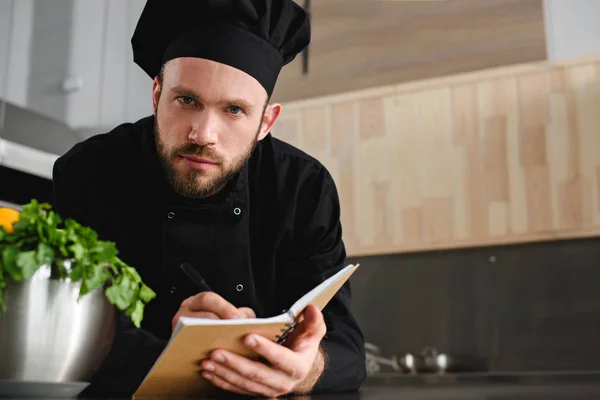 Guapo chef escribiendo una nueva receta para portátil en la cocina del restaurante y mirando a la cámara - foto de stock