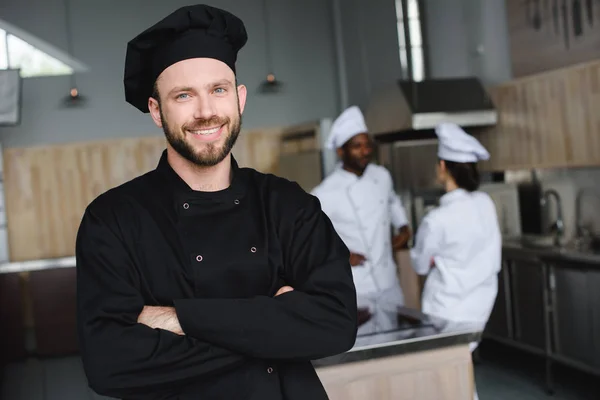 Улыбающийся шеф-повар стоит со скрещенными руками и смотрит в камеру на кухне ресторана — стоковое фото