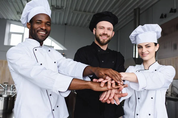 Chefs multiculturales sonrientes poniendo manos juntas en la cocina del restaurante - foto de stock