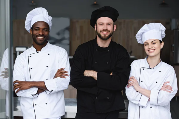 Усміхнені мультикультурні кухарі стоять з схрещеними руками і дивляться на камеру на кухні ресторану — стокове фото