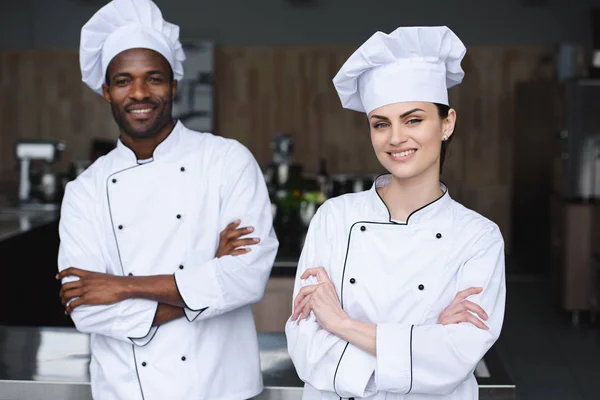 Multicultural chefs standing with crossed arms and looking at camera at restaurant kitchen — Stock Photo