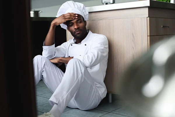 Exhausto chef afroamericano sentado en el piso en la cocina del restaurante y mirando hacia otro lado - foto de stock