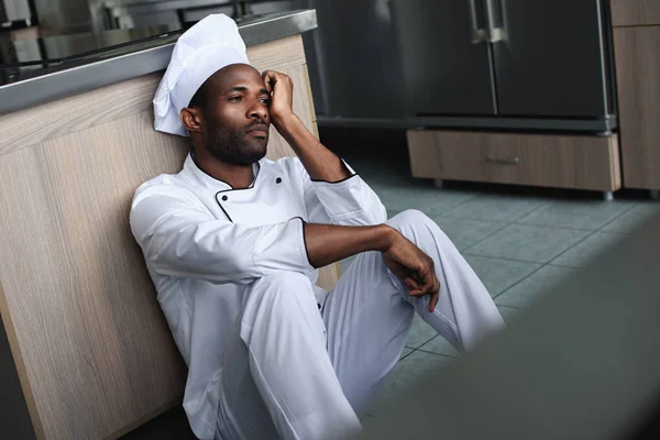 Chef afroamericano cansado sentado en el piso en la cocina del restaurante y mirando hacia otro lado - foto de stock