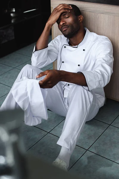 Exhausted african american chef sitting on floor at restaurant kitchen — Stock Photo