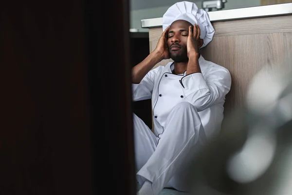 Chef africano cansado americano sentado no chão na cozinha do restaurante — Fotografia de Stock