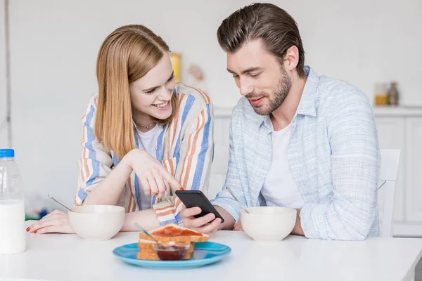 Lächelndes junges Paar nutzt Smartphone und sitzt mit Frühstück am Tisch — Stockfoto