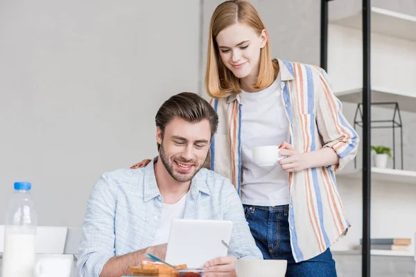 Atractiva mujer sosteniendo hombro novio mientras que el uso de tableta digital en la mesa con el desayuno - foto de stock