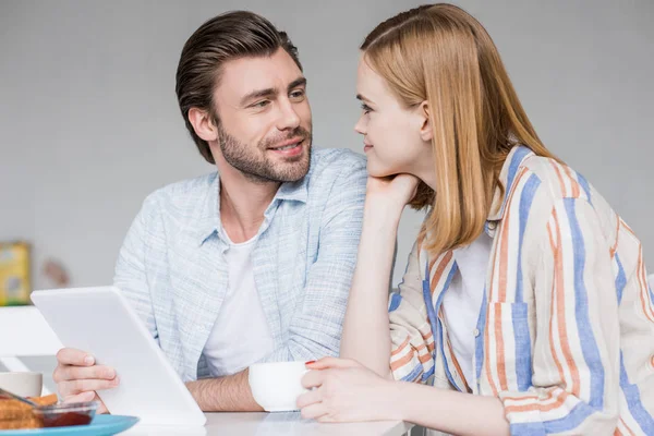 Atractiva joven con taza de café y novio con tableta digital mirándose entre sí - foto de stock