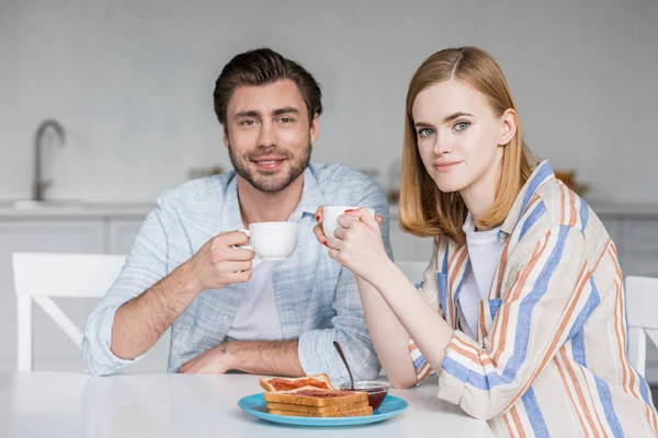 Sonriente pareja joven con tazas de café desayunando en la mesa en la cocina - foto de stock