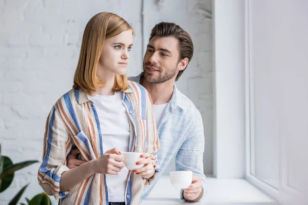 Stylish couple standing with coffee cups near window sill — Stock Photo
