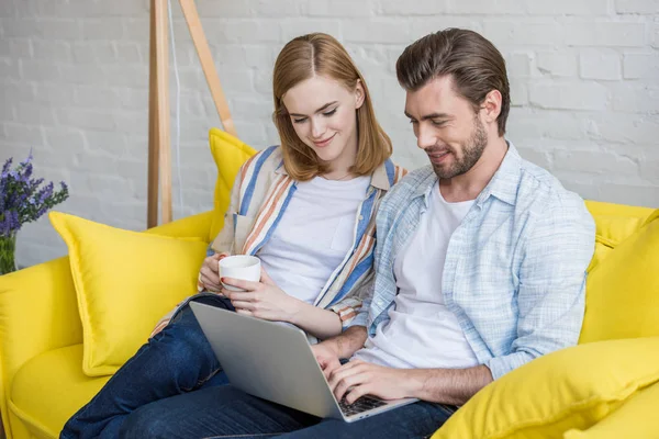 Homme souriant tapant sur ordinateur portable et petite amie avec tasse de café assis à côté — Photo de stock