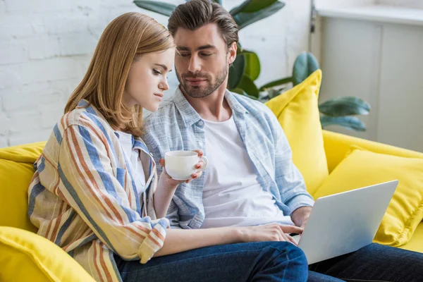 Young woman with coffee cup pointing by finger on laptop screen to boyfriend — Stock Photo
