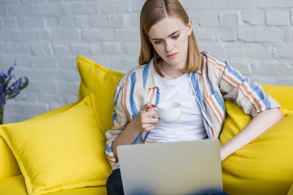 Mujer joven con taza de café usando el ordenador portátil y sentado en el sofá - foto de stock