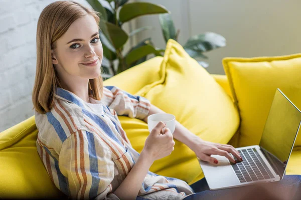 Улыбающаяся молодая женщина с чашкой кофе сидит на диване и с помощью ноутбука — стоковое фото