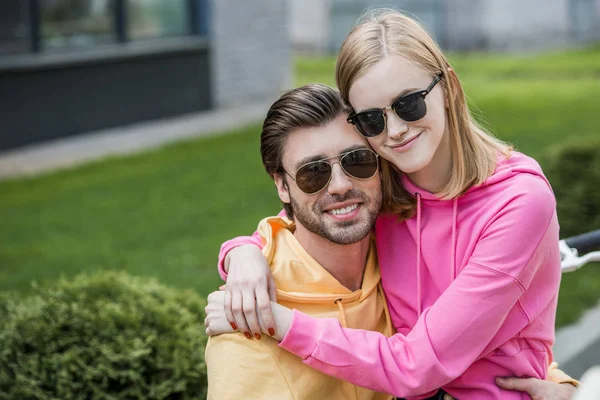 Smiling young woman in sunglasses sitting on boyfriend knees and embracing him — Stock Photo