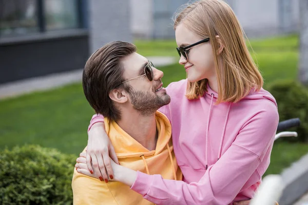 Femme élégante dans des lunettes de soleil embrassant petit ami et assis sur ses genoux — Photo de stock