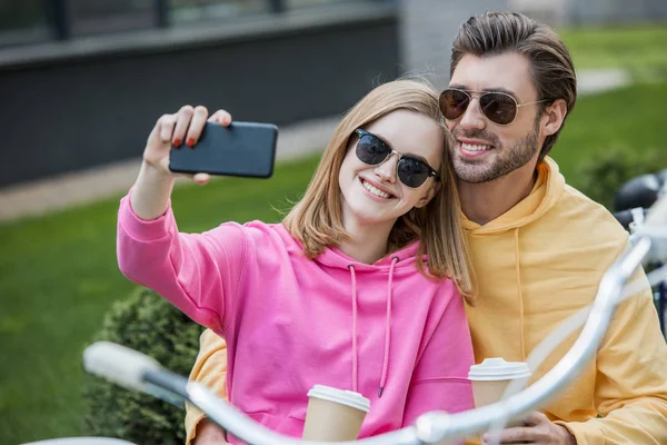 Sorrindo jovem mulher em óculos de sol tomando selfie com namorado — Fotografia de Stock