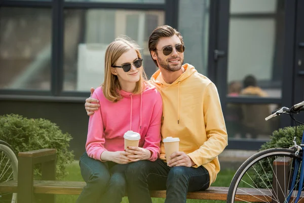 Lächelndes junges stilvolles Paar mit Sonnenbrille, Pappbecher mit Kaffee in der Hand und auf Bank sitzend — Stockfoto