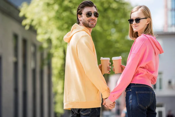 Elegante pareja en gafas de sol con tazas de café tomados de la mano uno del otro - foto de stock