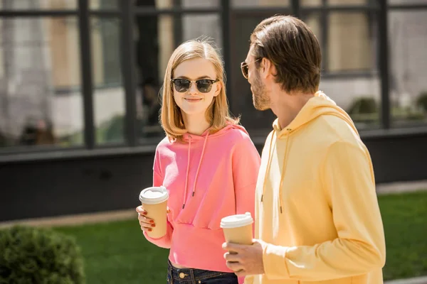 Jeune femme souriante dans des lunettes de soleil avec tasse de café marche avec petit ami dans la rue — Photo de stock