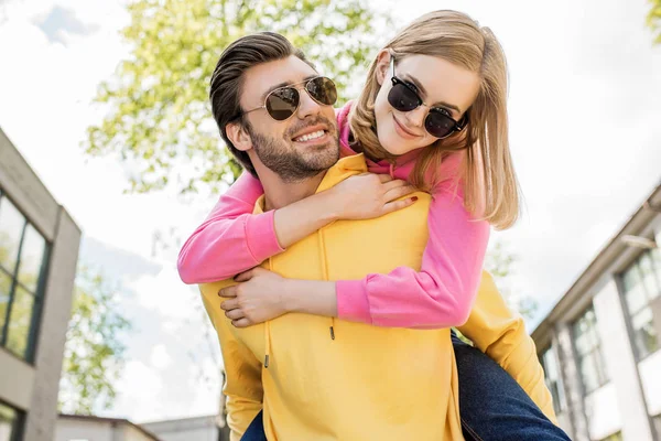 Jovem homem em óculos de sol fazendo passeio de piggyback para namorada — Fotografia de Stock