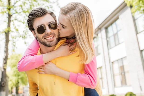 Stylish man in sunglasses doing piggyback ride to girlfriend while she kissing him in cheek — Stock Photo
