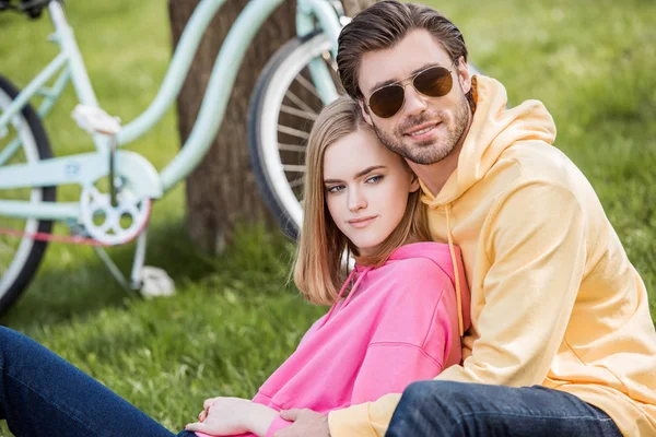 Joven con estilo en gafas de sol sentado en la hierba con la novia - foto de stock