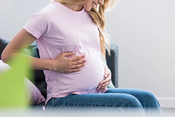 Imagen recortada de la mujer embarazada sonriente sentada en el sofá y tocando el vientre - foto de stock