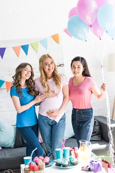 Amigos multiculturales sonrientes y mujer embarazada de pie con globos en la fiesta del bebé - foto de stock