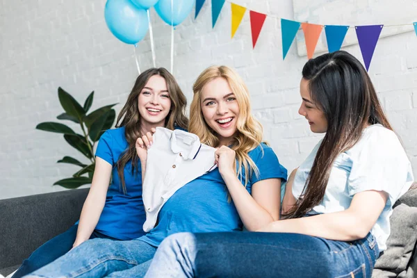 Happy pregnant woman showing baby clothes and sitting with multicultural friends at baby-party — Stock Photo